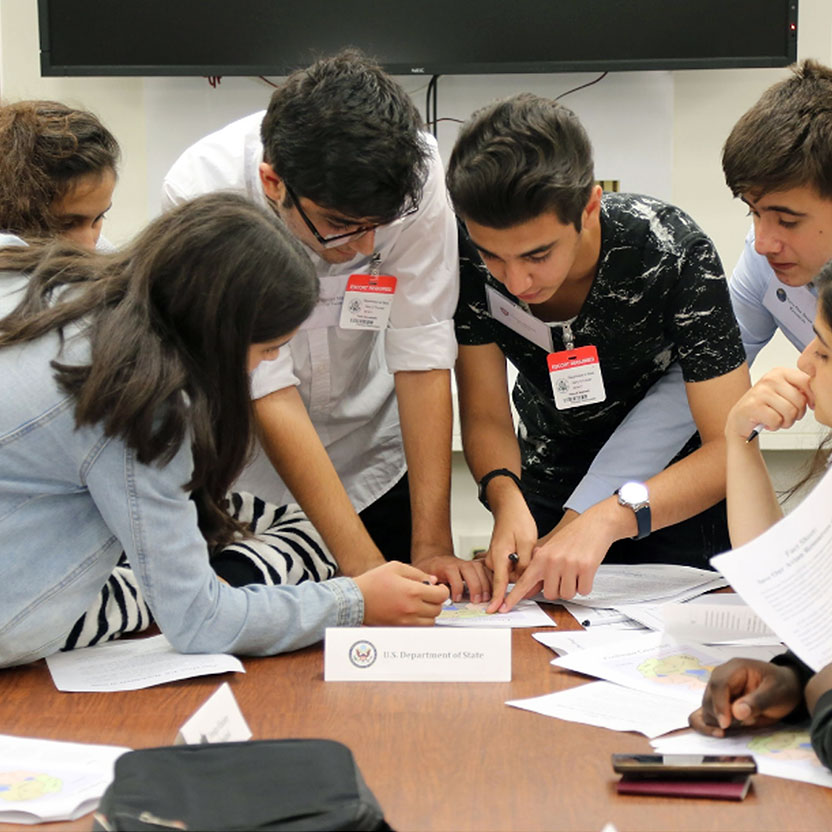 A group of students around a table looking at papers, pointing, and discussing.